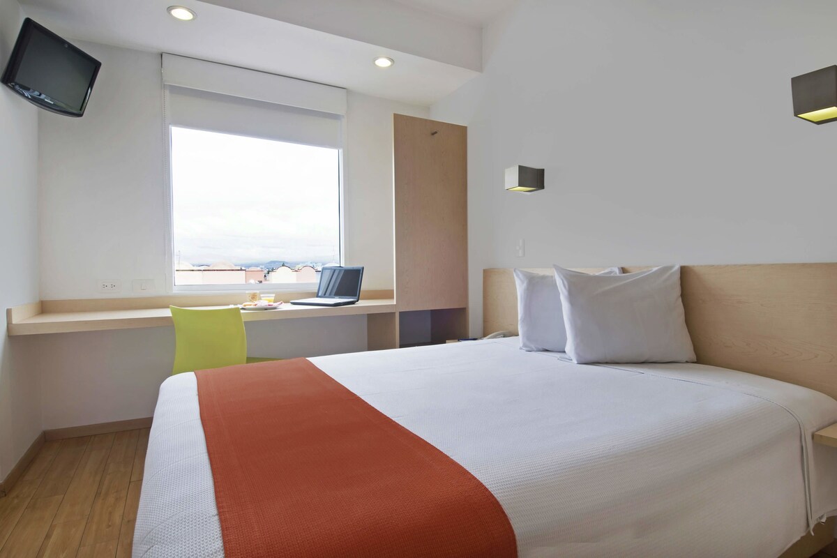 A hotel room with a bed, a desk with a laptop, a TV mounted on the wall, and a window with a view.