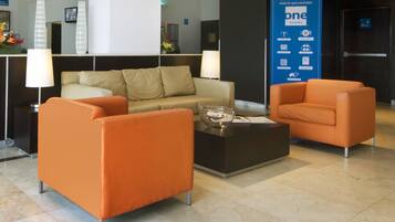 A hotel lobby with orange armchairs, a black coffee table, and a beige sofa. A large blue sign with hotel amenities is visible.