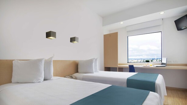 Een hotelkamer met twee bedden, een groot raam met uitzicht over de stad, een bureau met een laptop en een wandgemonteerde flatscreen-tv.