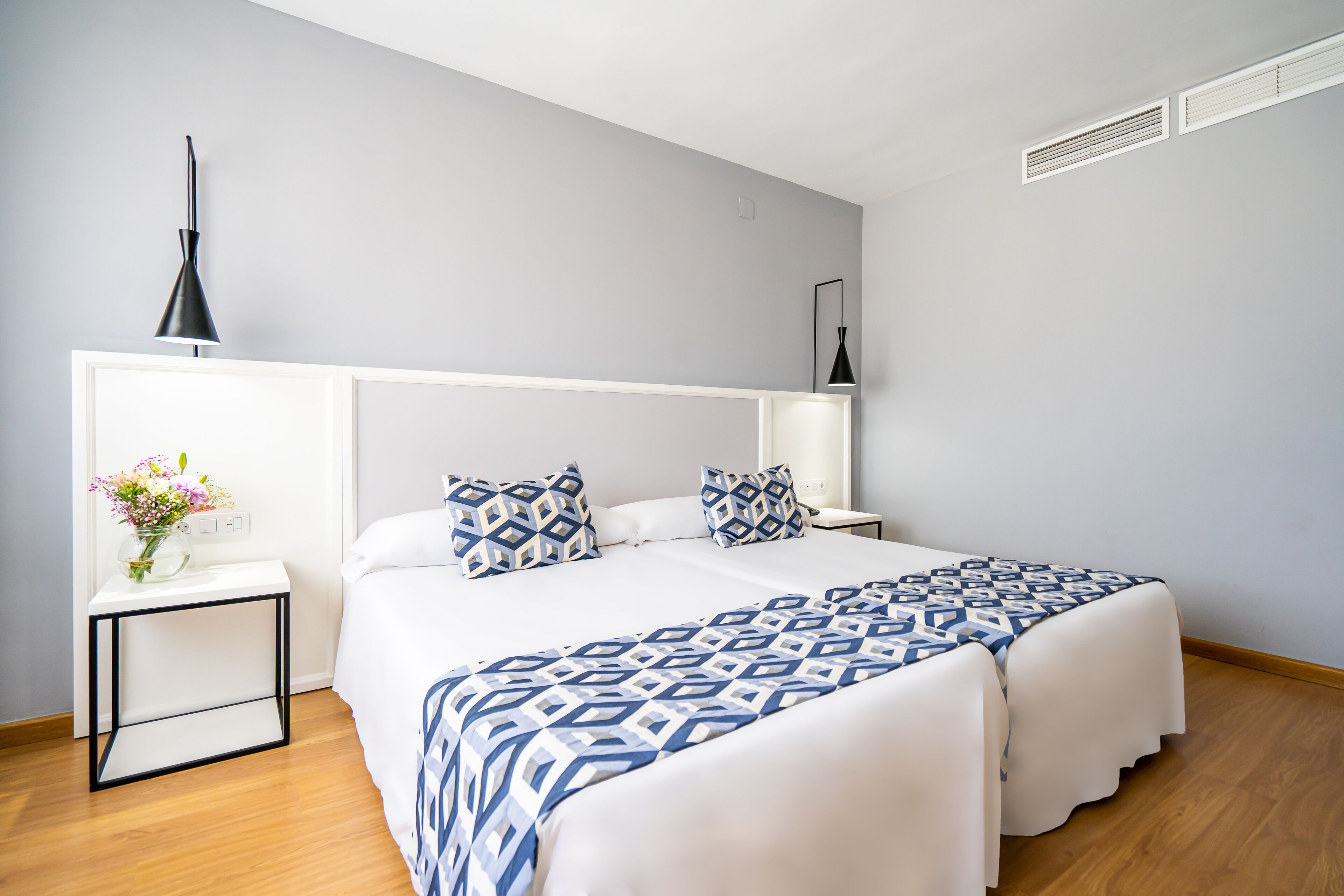 A modern hotel room with a large bed, bedside tables, a vase with flowers, and a ceiling vent.
