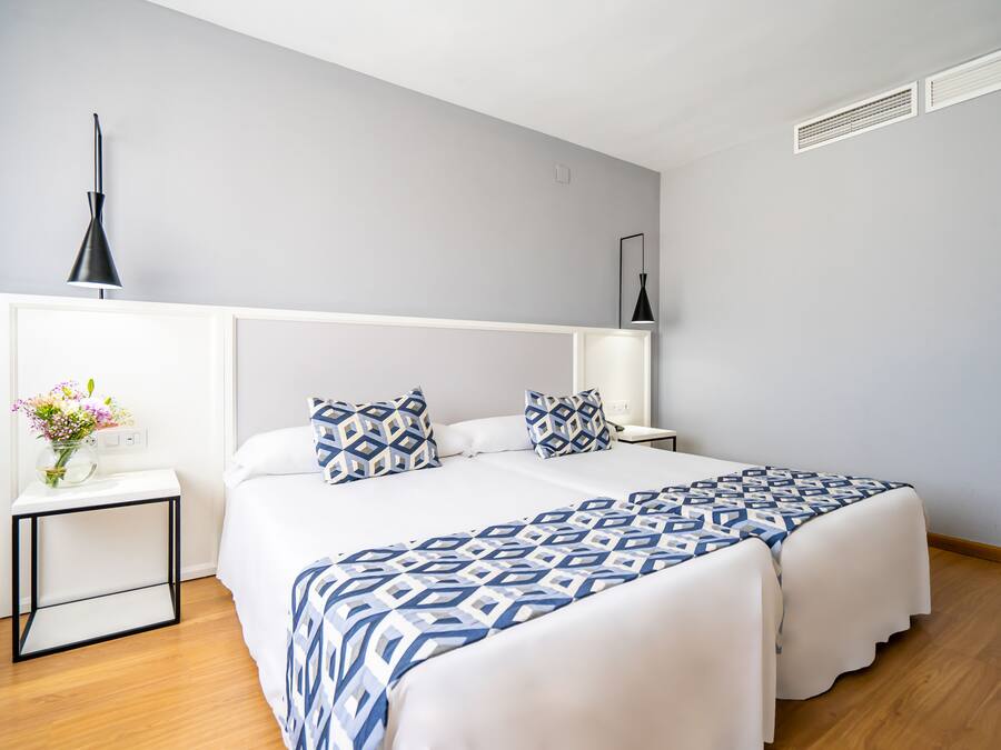 Una habitación de hotel moderna con una cama grande, mesitas de noche, un jarrón con flores y un respiradero en el techo.