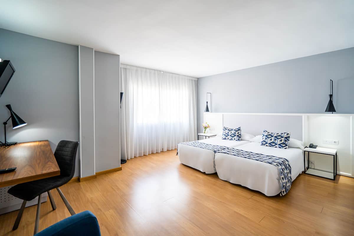A modern hotel room with a wooden floor, a desk with a chair, a bed with patterned bedding, and a bedside table with a lamp.