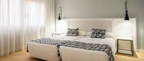 A double bed with a geometric-patterned duvet, two bedside tables with black lamps, and a large window with sheer curtains.