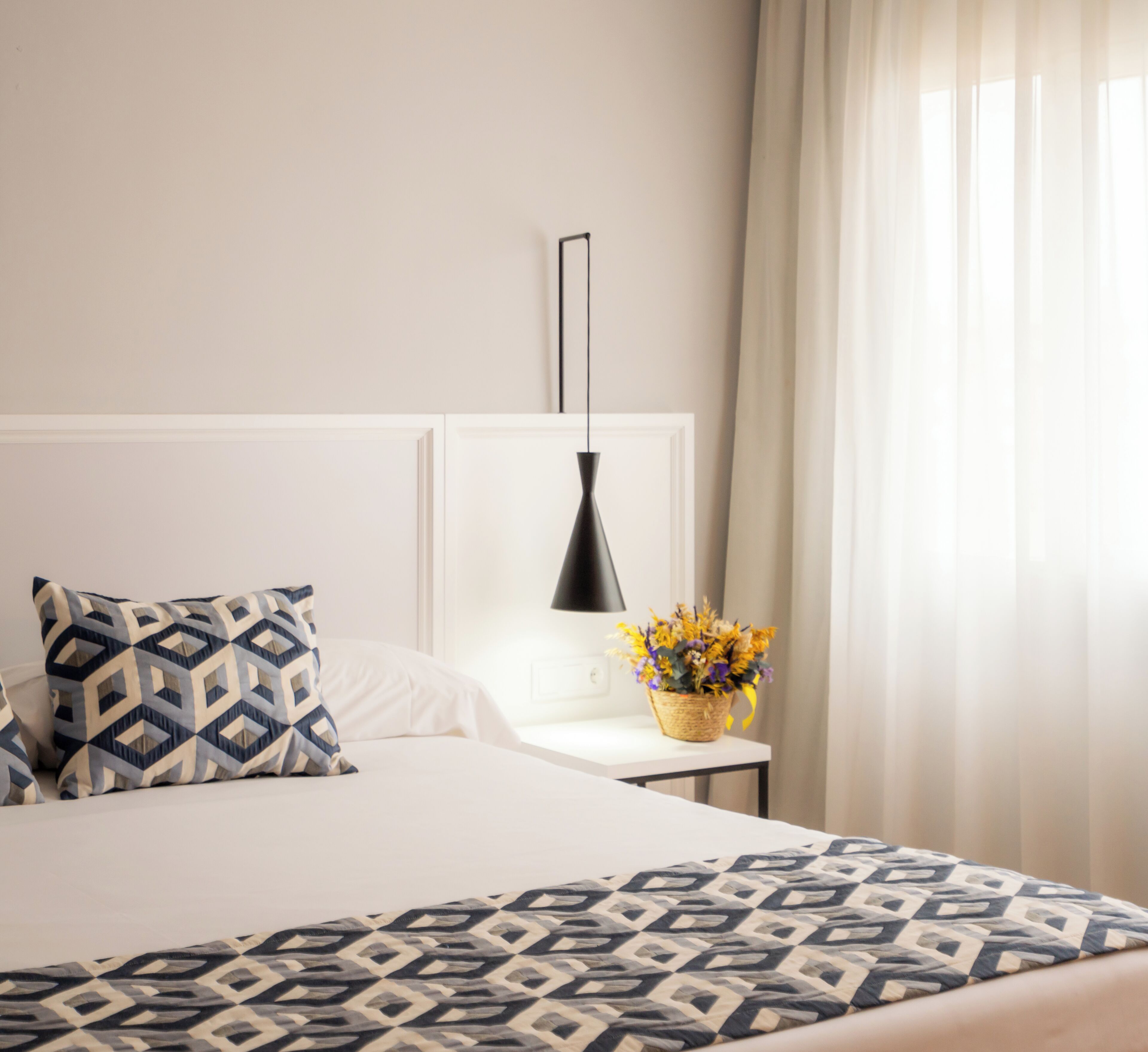 Un letto ben rifatto con cuscini decorati, un comodino con una lampada e un vaso di fiori, e una finestra con tende.