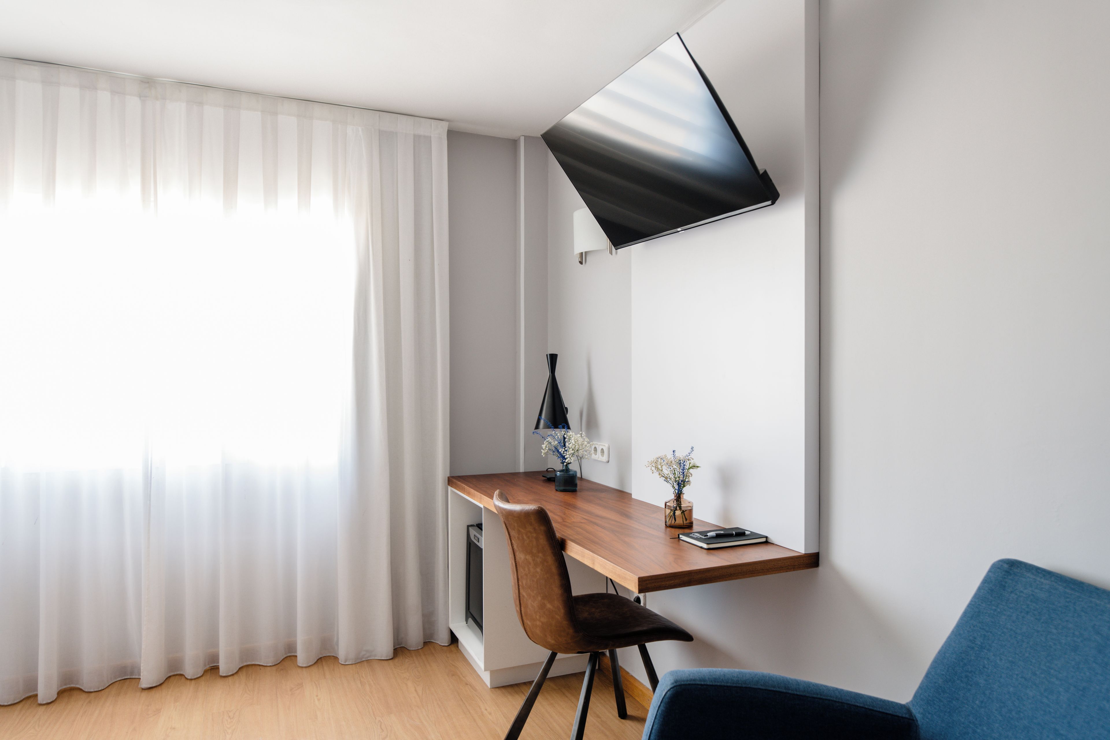 Habitació moderna amb escriptori de fusta, una cadira, televisió plana penjada a la paret i finestra amb cortines primes.