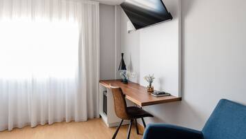 Habitació moderna amb escriptori de fusta, una cadira, televisió plana penjada a la paret i finestra amb cortines primes.