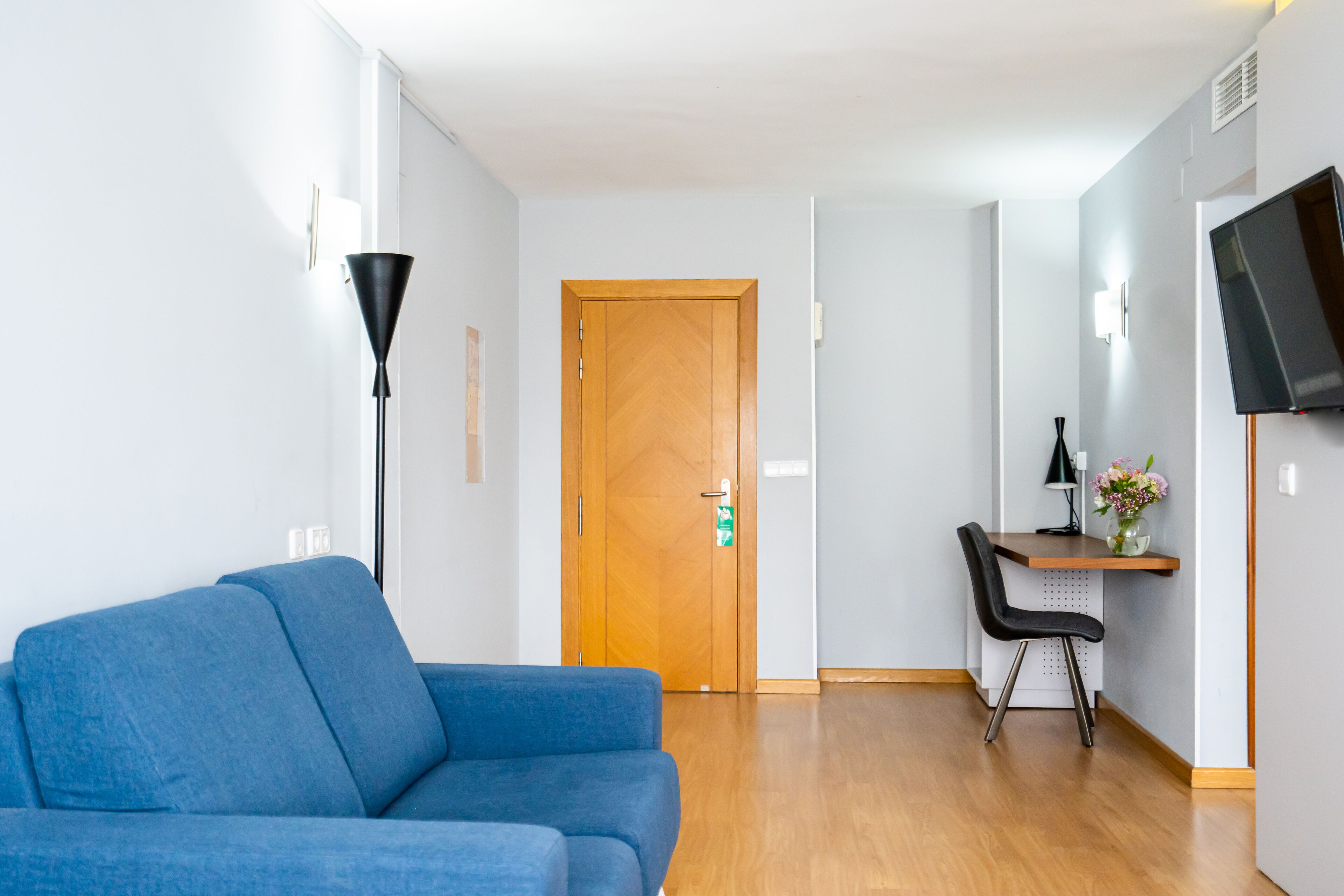 Una camera d'albergo moderna con un divano blu, una scrivania con una sedia, un televisore montato a parete e una porta in legno.