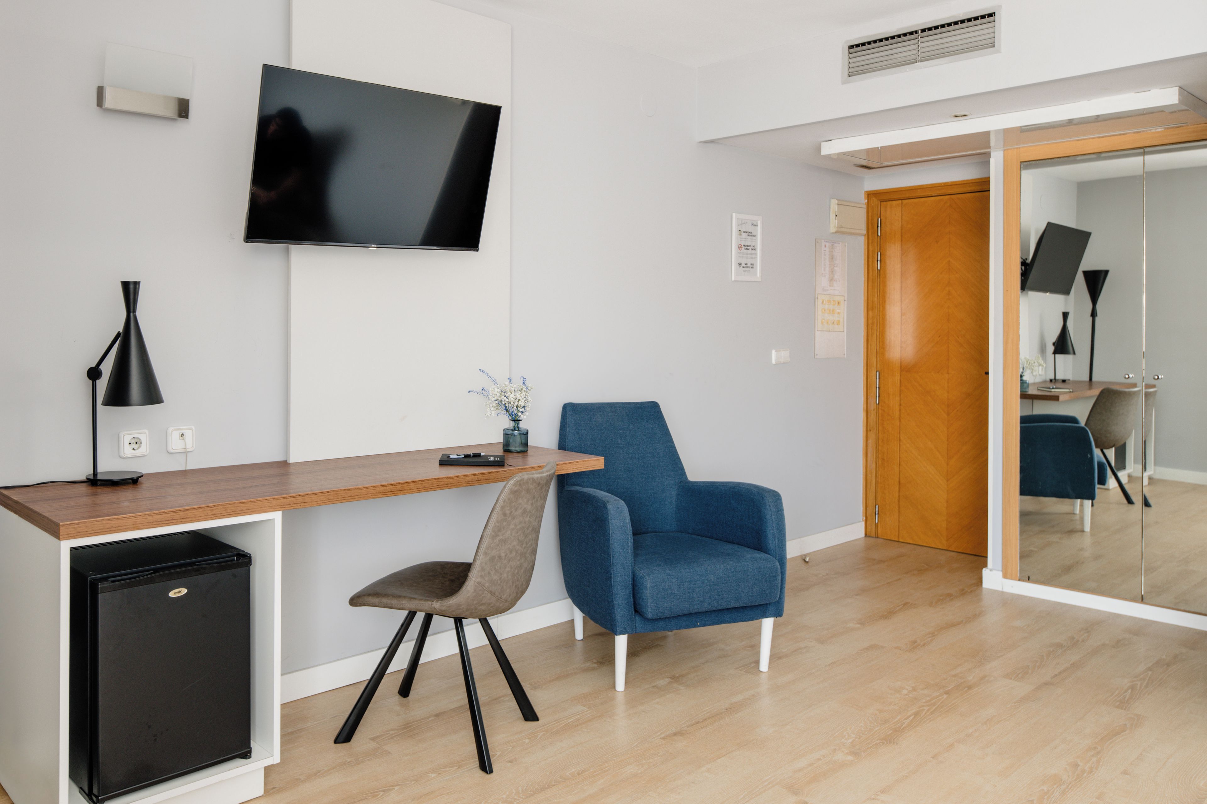 A modern room with a wooden desk, a blue chair, a small black fridge, and a flat-screen TV mounted on the wall.