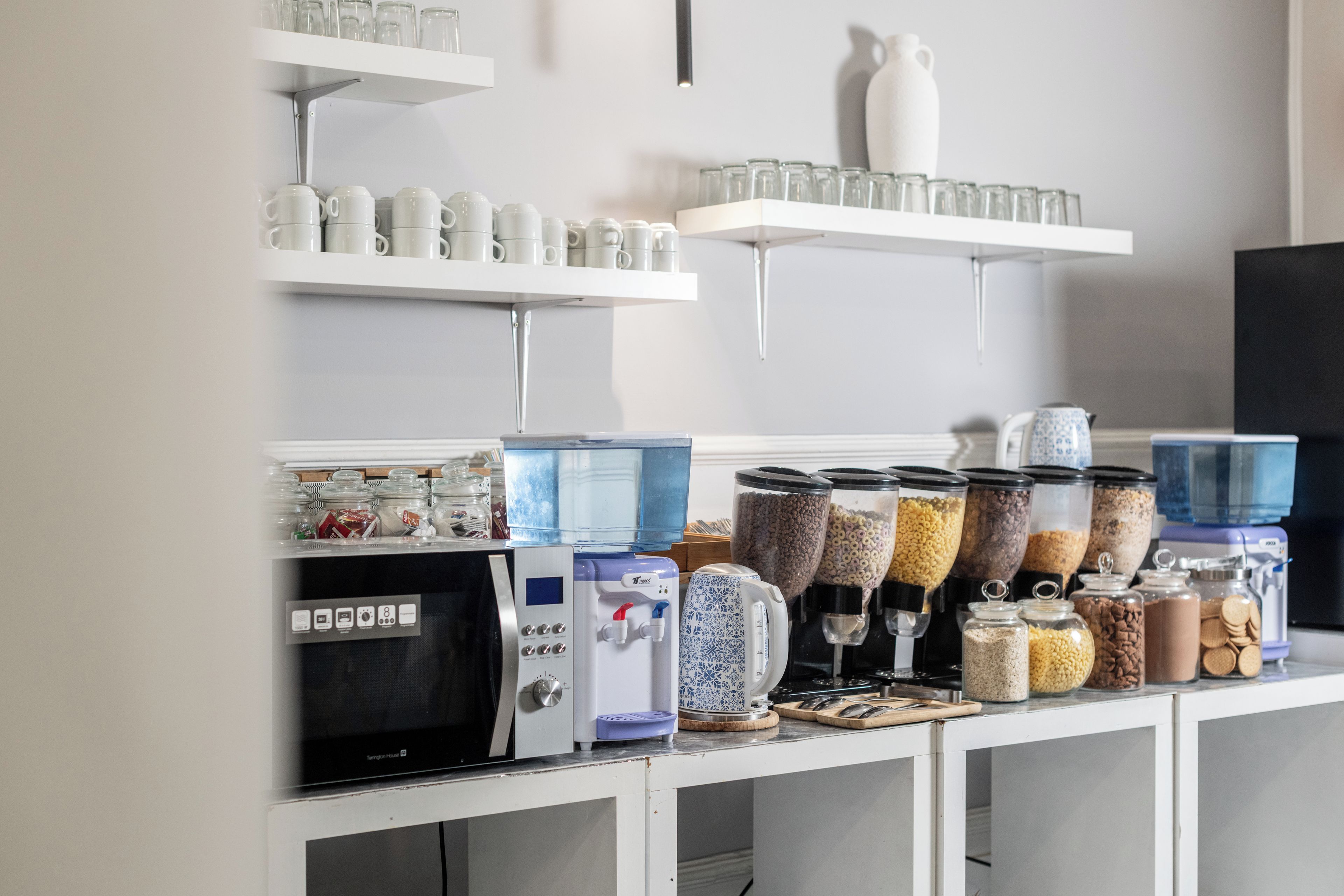 Un área de cocina con estantes que sostienen tazas y vasos, un dispensador de agua y varios recipientes para almacenar alimentos.