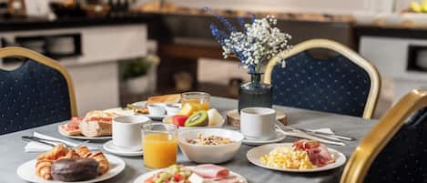 Una mesa de desayuno con diversos alimentos y bebidas, un florero con flores y una cocina al fondo.