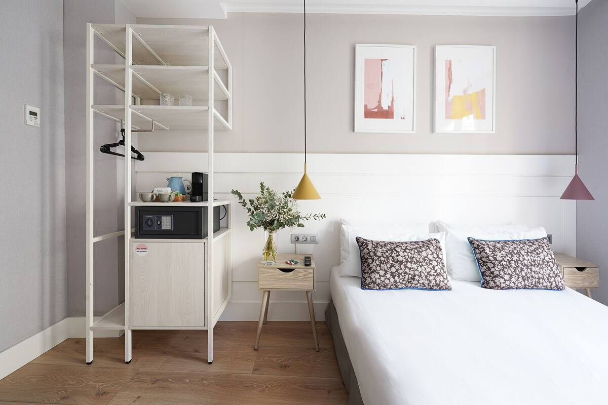 A modern bedroom with a bed, two pillows, a nightstand, a coffee maker, a kettle, a vase with flowers, a wardrobe, and two wall art pieces.