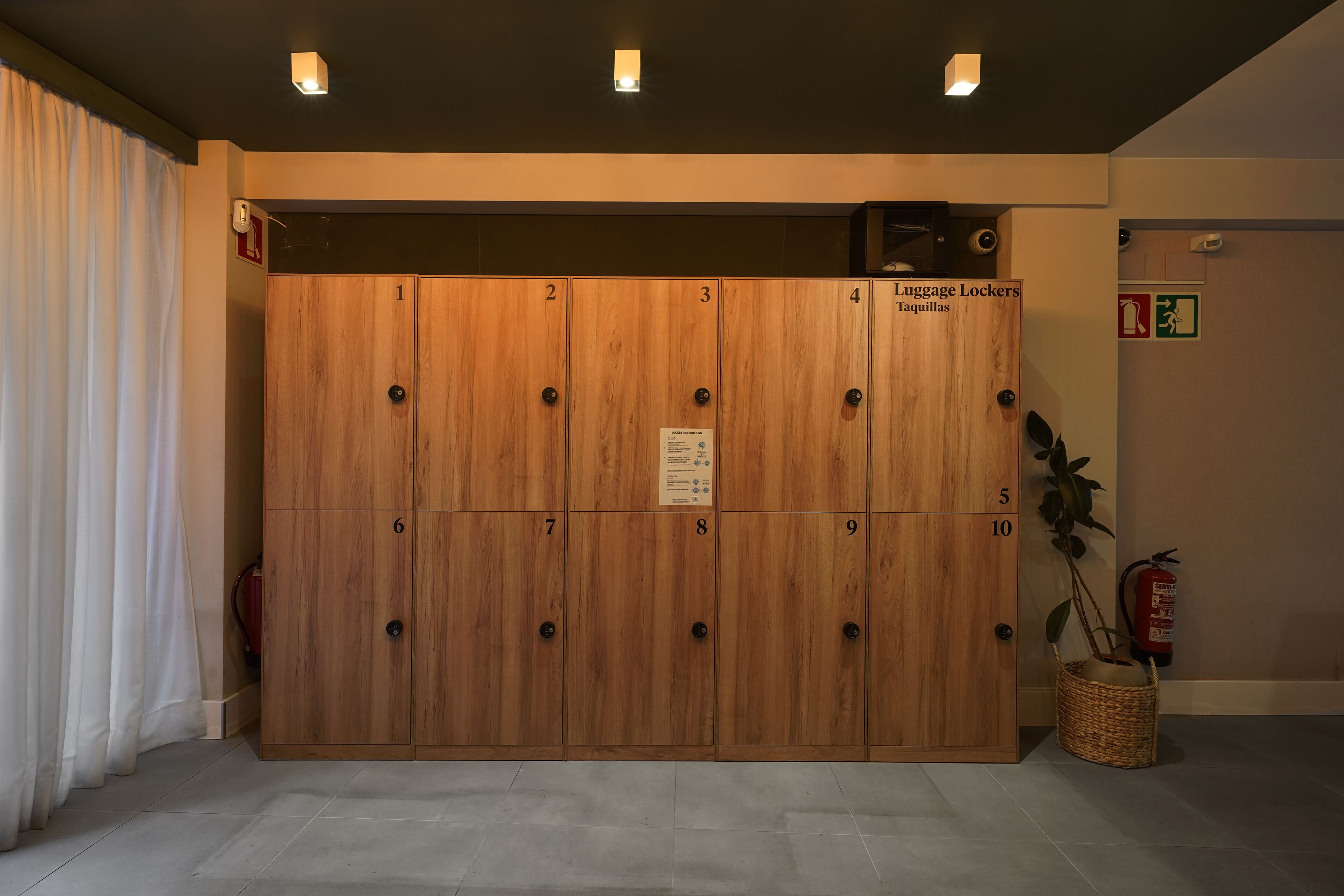 A row of wooden lockers with numbered doors, a sign indicating 'Luggage Lockers Taquillas', and a fire extinguisher mounted on the wall.