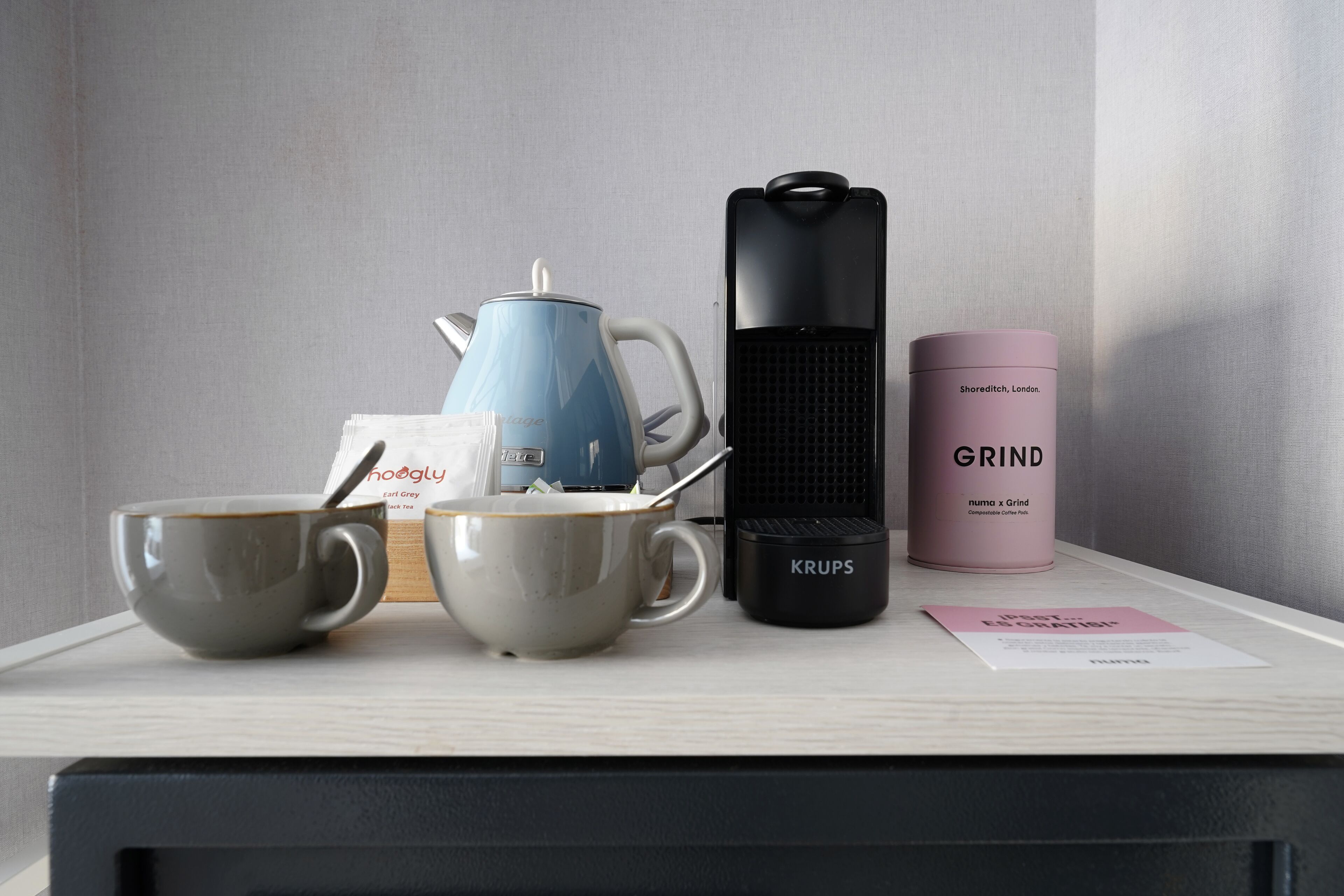 Un ensemble pour préparer le café comprenant une bouilloire bleue, deux tasses, une machine à café Krups et un récipient rose portant l’inscription « MOUTURE ».