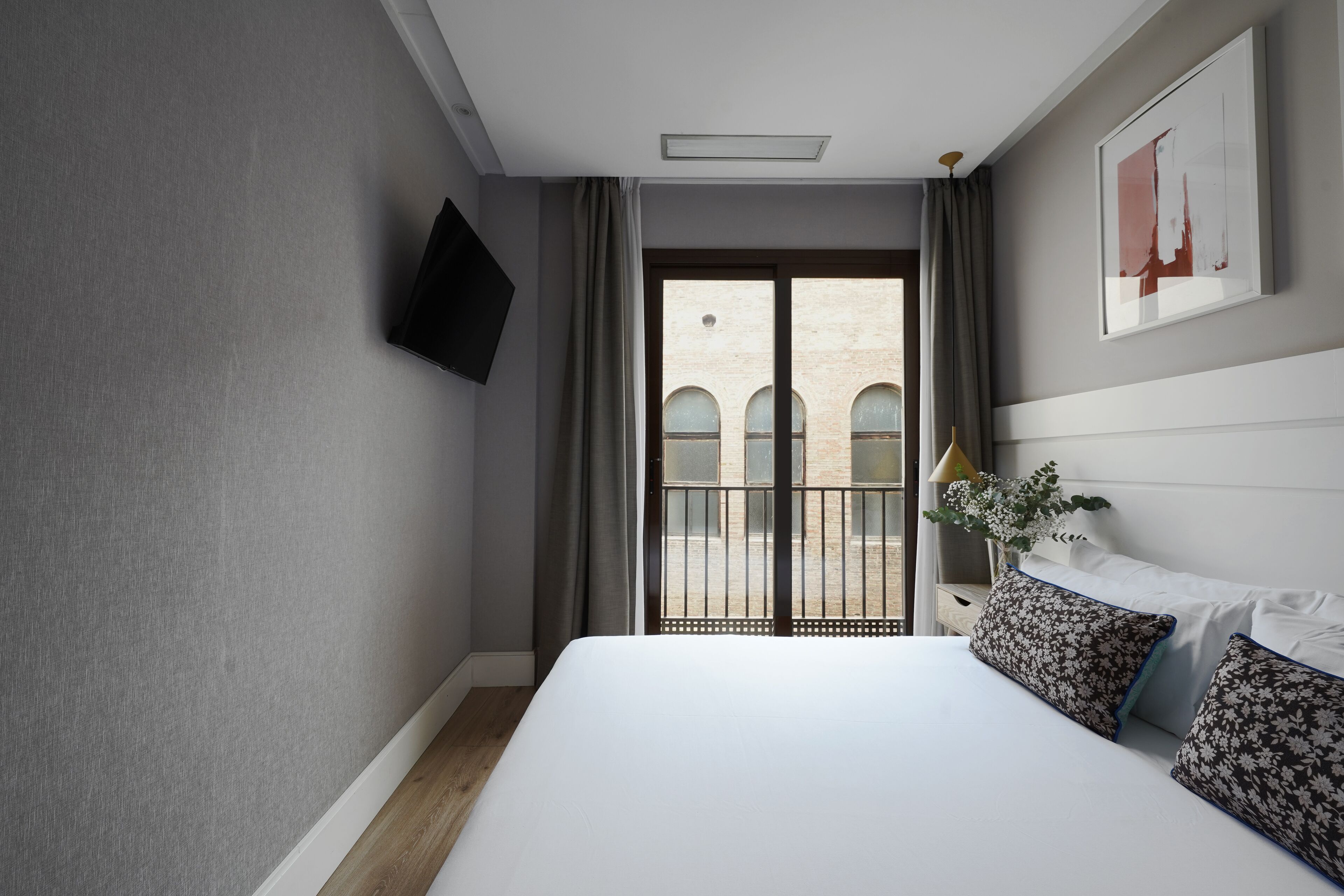 A hotel room with a large bed, a flat-screen TV mounted on the wall, a balcony with a view of a building, and a framed picture on the wall.