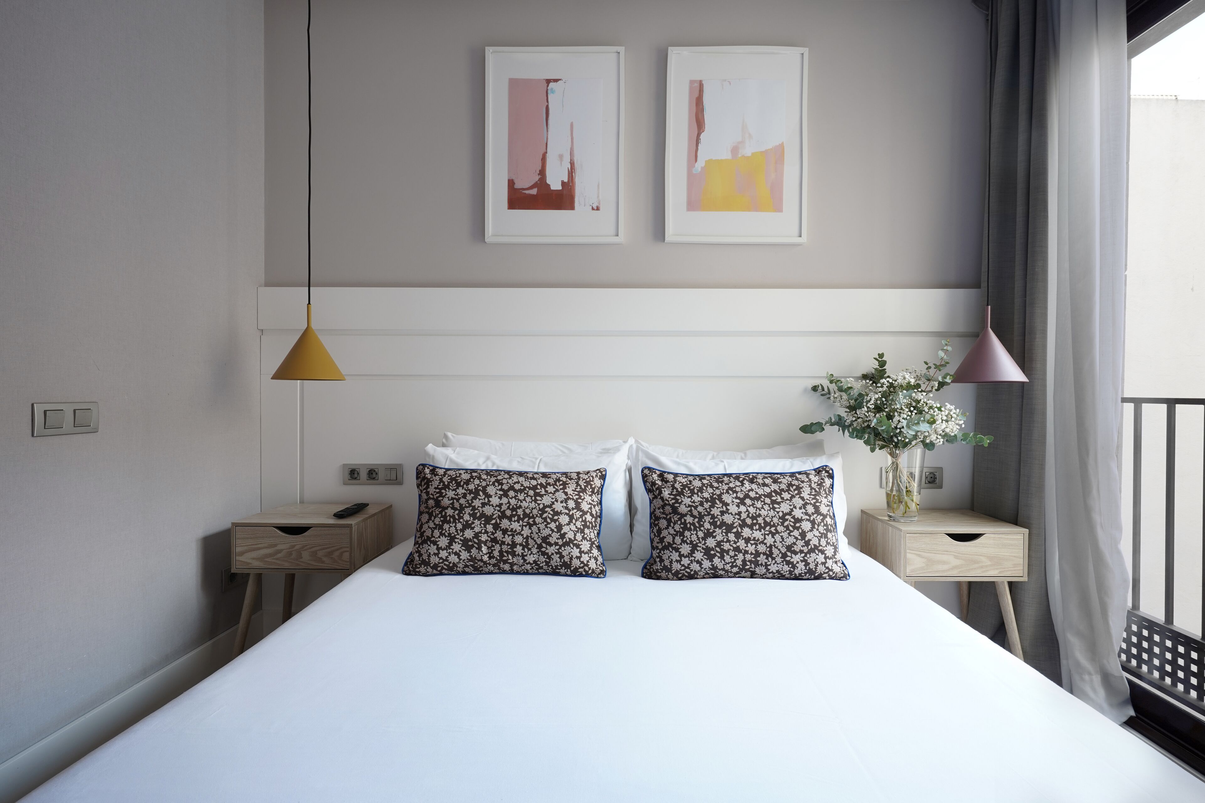 A modern hotel room with a large bed, two bedside tables, a nightstand, a vase with flowers, and two abstract paintings on the wall.