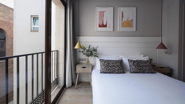 A modern bedroom with a large bed, a nightstand, a small table, and a window with curtains. There are two framed artworks on the wall above the bed. - Numa Barcelona Brio (Barcelona)