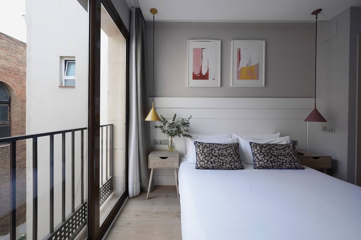 A modern bedroom with a large bed, a nightstand, a small table, and a window with curtains. There are two framed artworks on the wall above the bed.