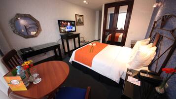A hotel room with a bed, a desk with a television, a dining table with chairs, and a vase with flowers.