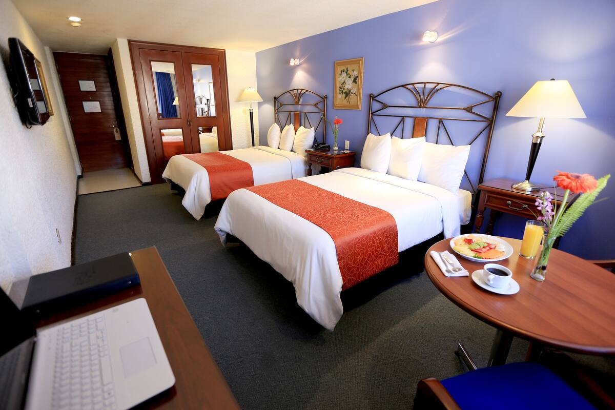 Ein Hotelzimmer mit zwei Betten, einem Schreibtisch mit einem Laptop, einem Esstisch mit Frühstück und einer Badezimmertür.