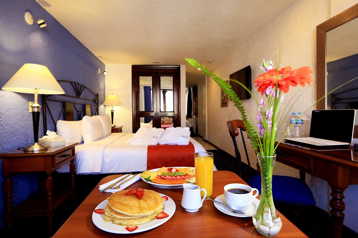 Ein Hotelzimmer mit Bett, Nachttisch mit Lampe, Esstisch mit Frühstücksutensilien, einer Vase mit Blumen und einem Schreibtisch mit Laptop.