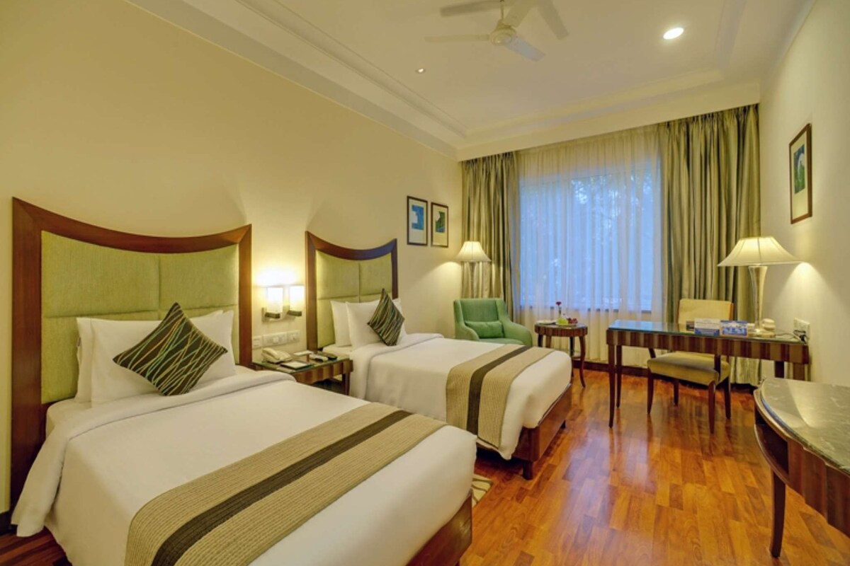 A hotel room with two beds, a desk, a chair, a lamp, a ceiling fan, and a window with curtains.