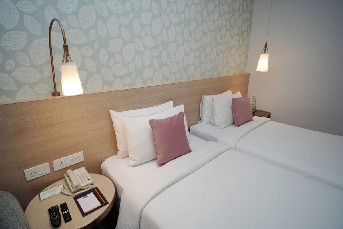 A hotel room with two beds, a bedside table with a phone and remote controls, a wall with a leaf pattern, and a hanging light fixture.