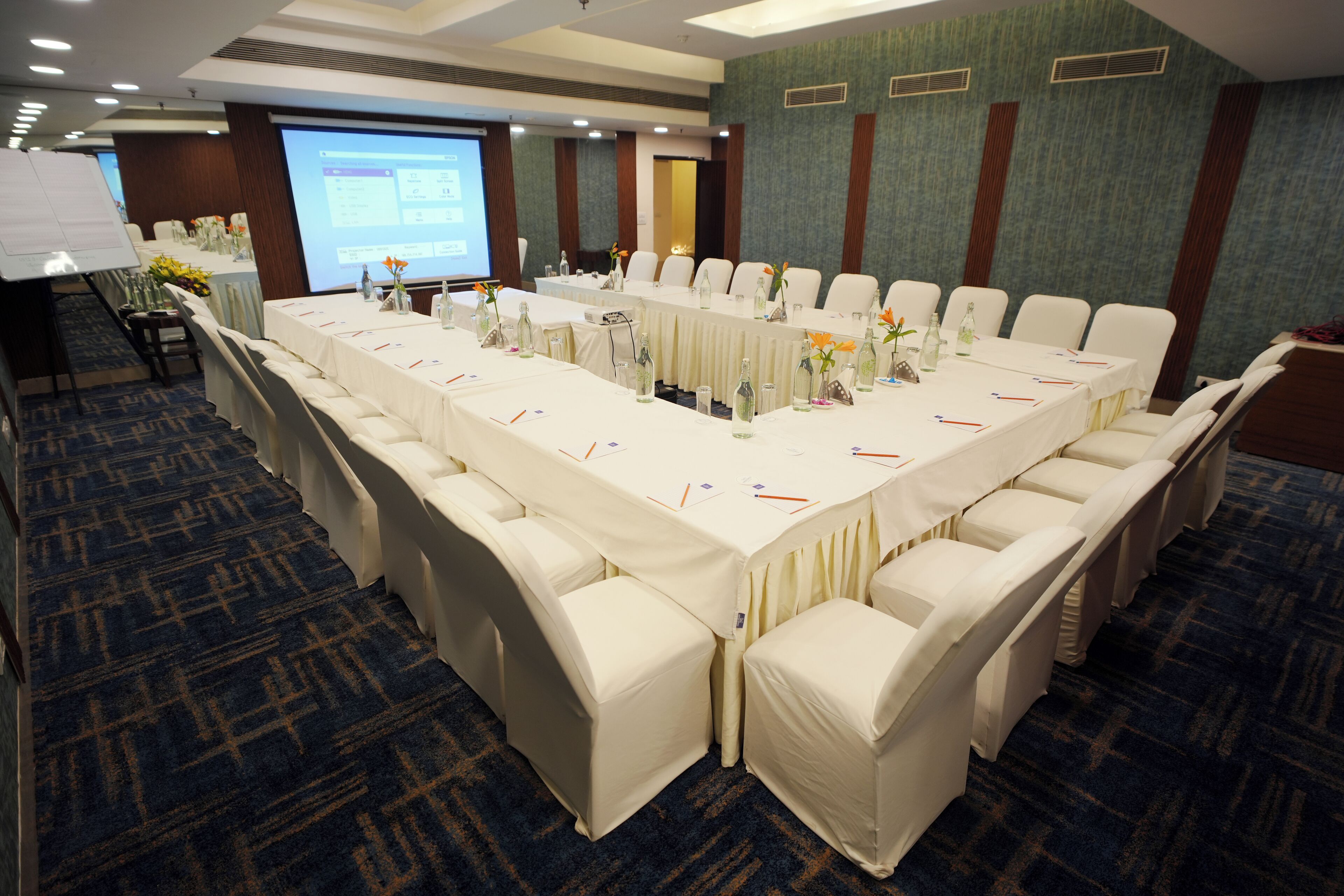 Un salón de conferencias con una larga mesa, sillas blancas y una pantalla para proyector.