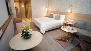 A modern hotel room with a large bed, a sofa, a small table, and a potted plant.