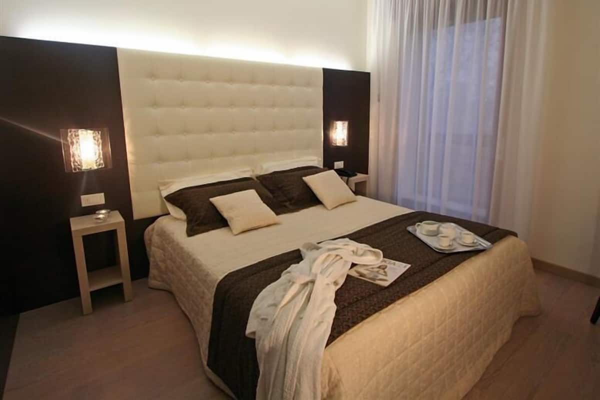 A hotel room with a large bed, two bedside tables, a nightstand, a window with curtains, and a wall-mounted light fixture.