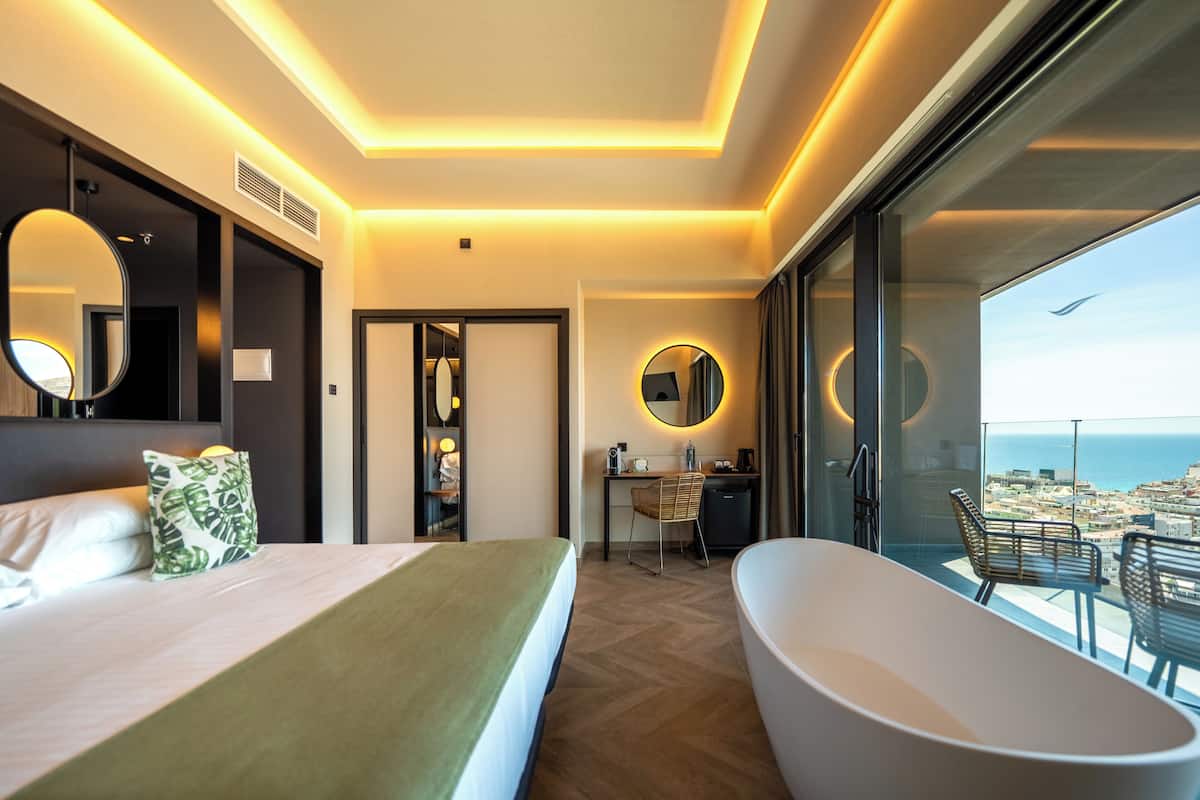 A modern hotel room with a large bed, a bathtub, a desk, and a view of the city.