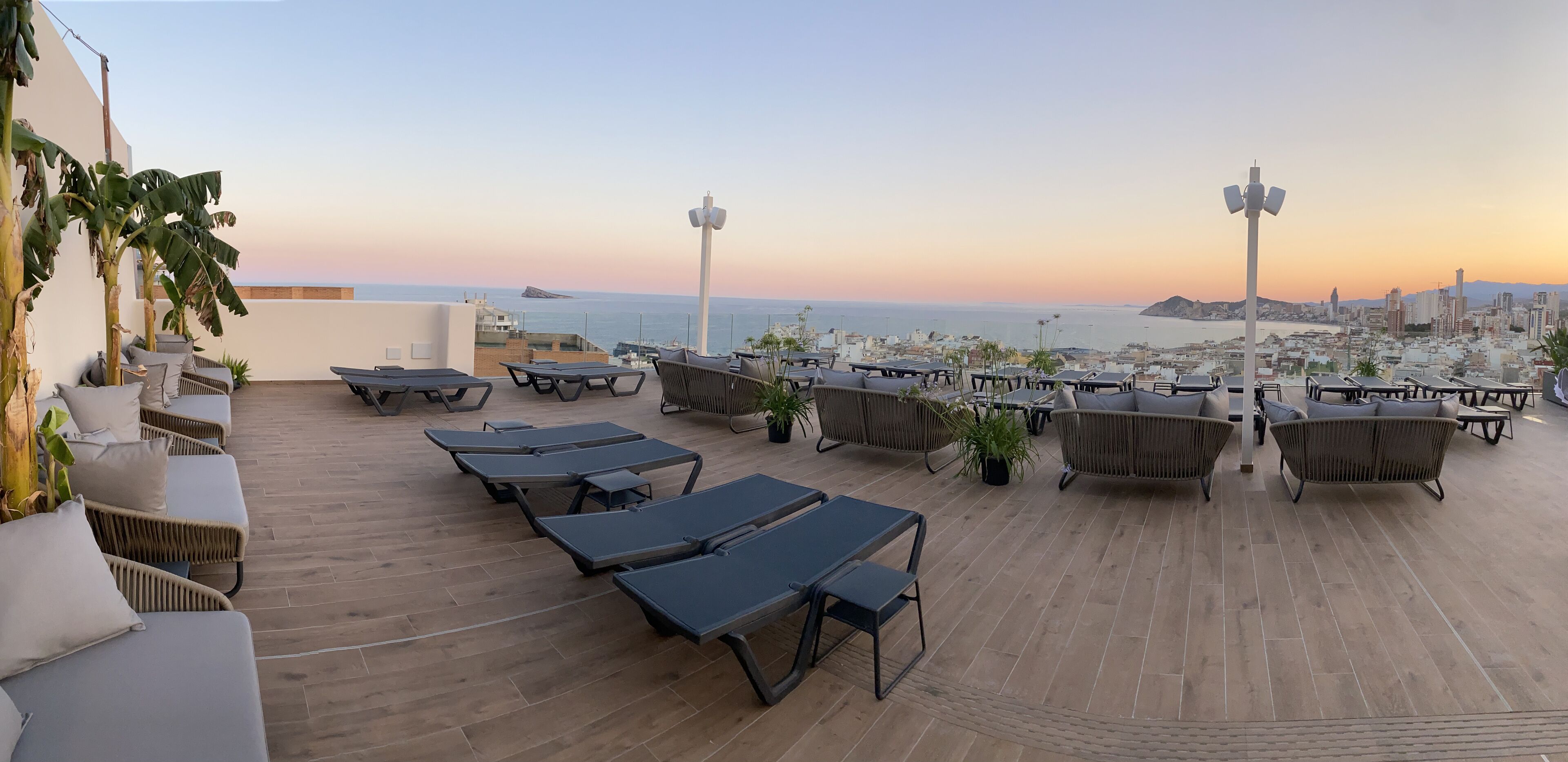 A rooftop terrace with lounge chairs, tables, and potted plants, overlooking a cityscape and the sea at sunset.