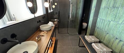 A modern bathroom with a large sink, mirror, and towel rack. There is a bench with towels and a small table with toiletries.