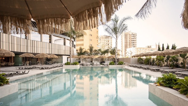Piscina con sillas de descanso y sombrillas, rodeada de edificios y palmeras.