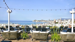Terraza en la azotea con sillones de mimbre y plantas en macetas, con vistas a un paisaje urbano costero.