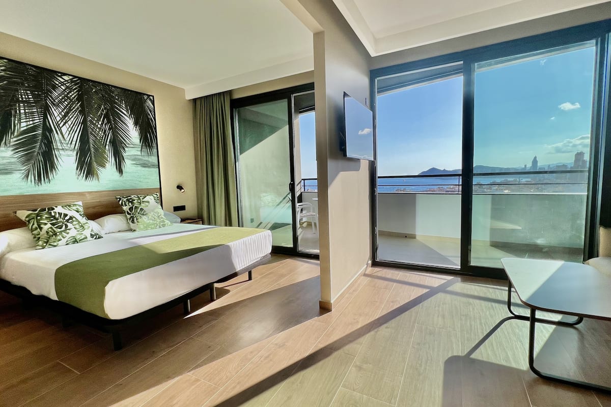 A modern hotel room with a bed, a TV, a balcony with a city view, and a dining area.