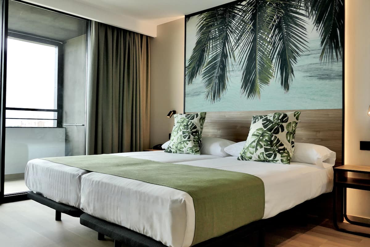 A modern hotel room with a large bed, two pillows with a tropical leaf pattern, a green throw blanket, a nightstand, and a wall-mounted lamp.