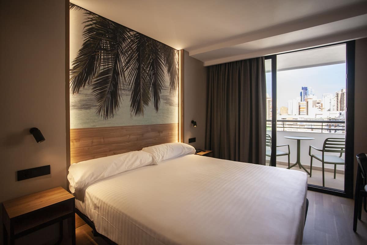 A hotel room with a large bed, a nightstand, a wall-mounted light, and a view of the city through a large window.