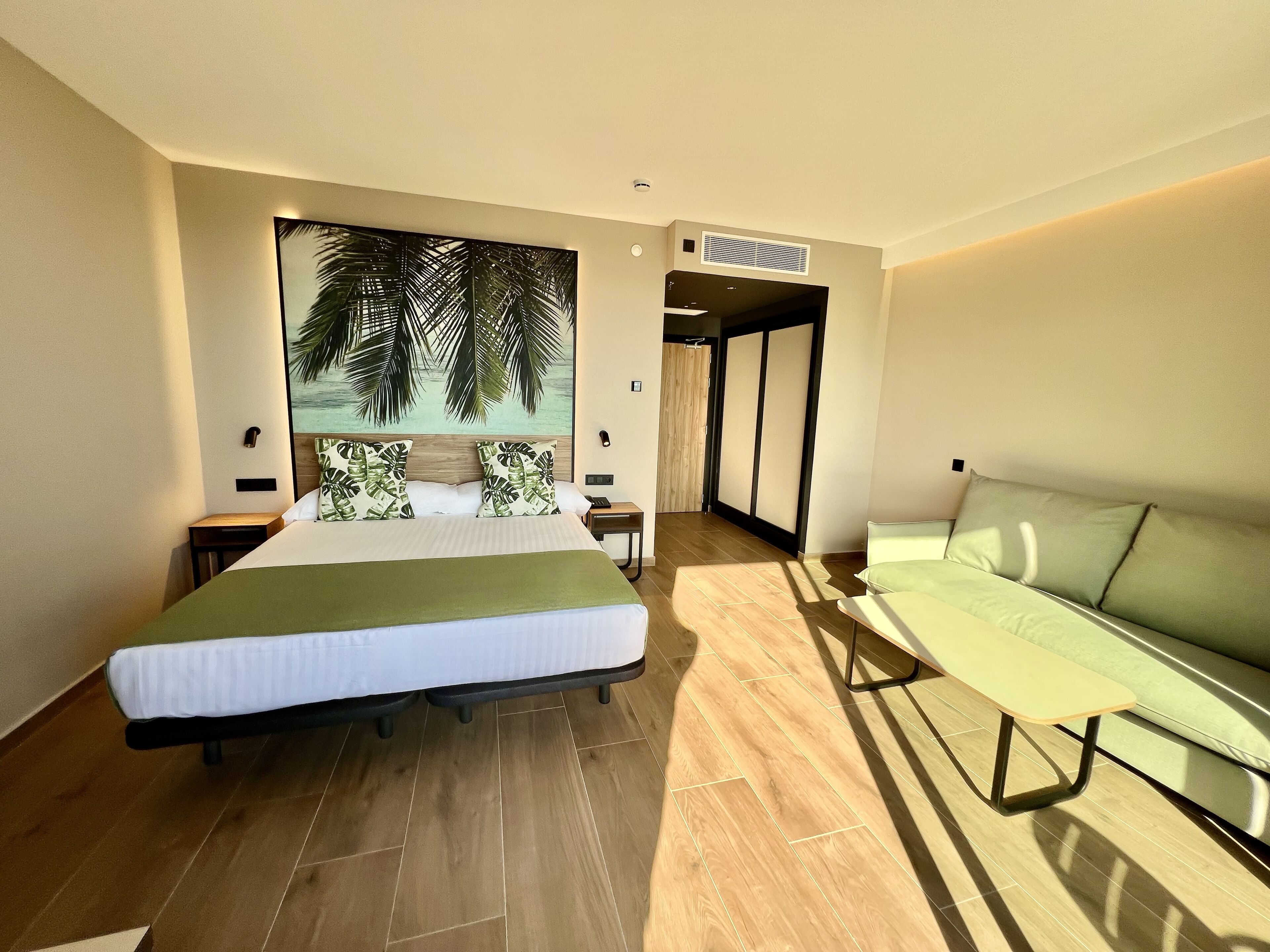 Ein Hotelzimmer mit einem Bett, einer grünen Schlafsofa, einem großen Wandbild, Holzboden und einer Schiebetür.