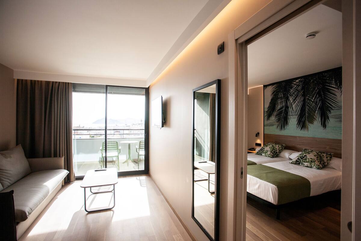 A modern hotel room with a balcony, a sofa, a bed with a green bedspread, and a large mirror.