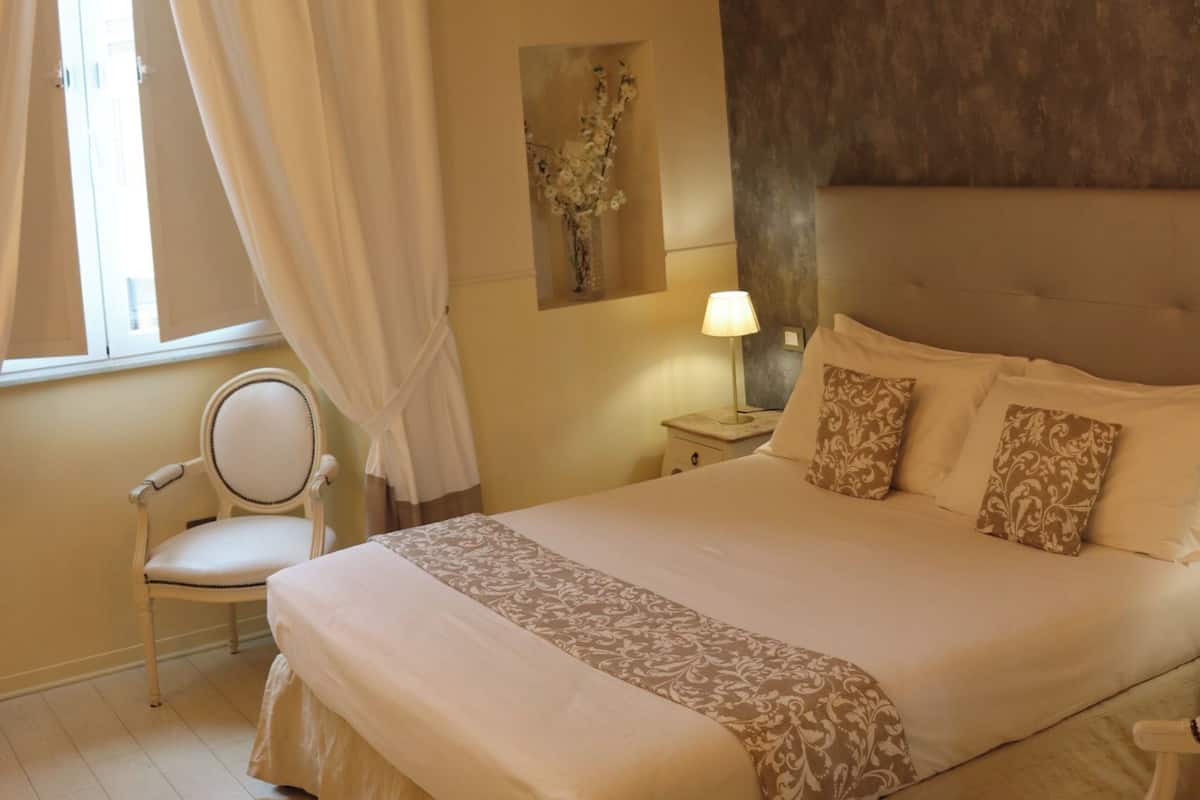 A bedroom with a large bed, a bedside table with a lamp, a chair, and a window with curtains.