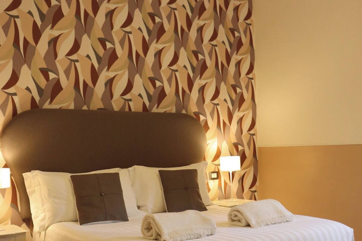 A neatly made bed with a headboard, two bedside lamps, and a patterned wall behind it.