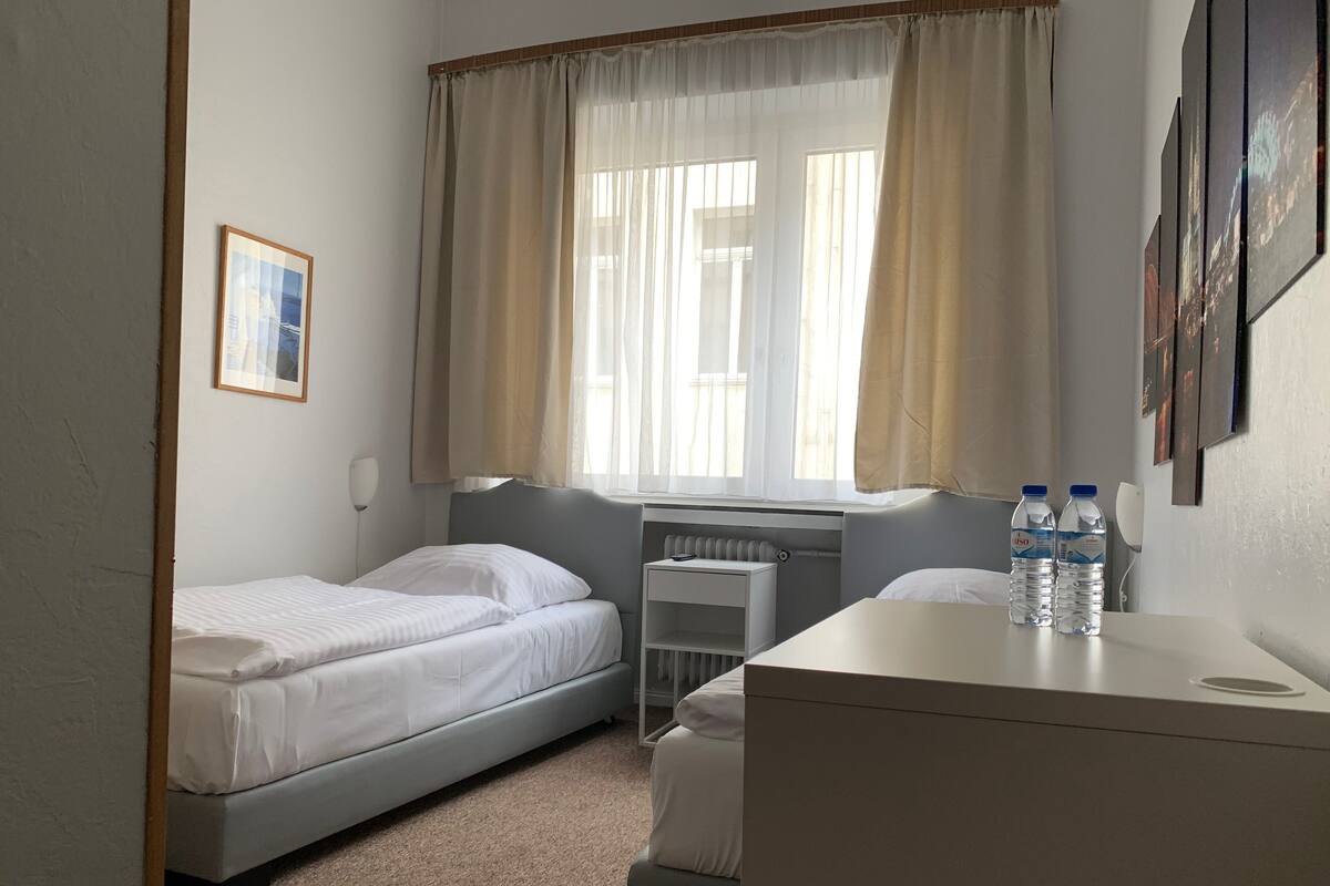 Habitación de hotel con dos camas individuales, un escritorio con una silla, un radiador y dos botellas de agua sobre el escritorio.