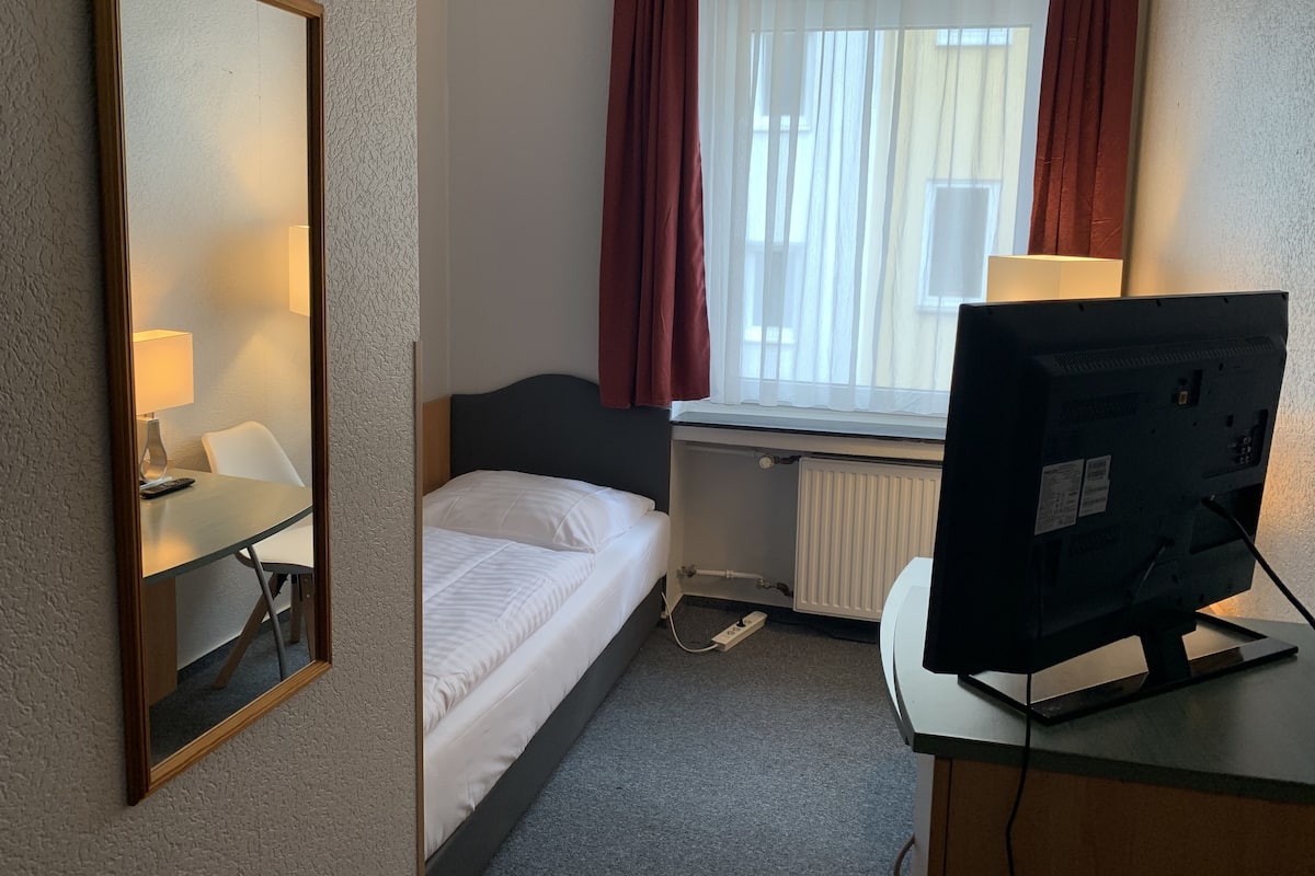 Habitación de hotel con una cama individual, un escritorio con ordenador, un espejo y una ventana con cortinas.