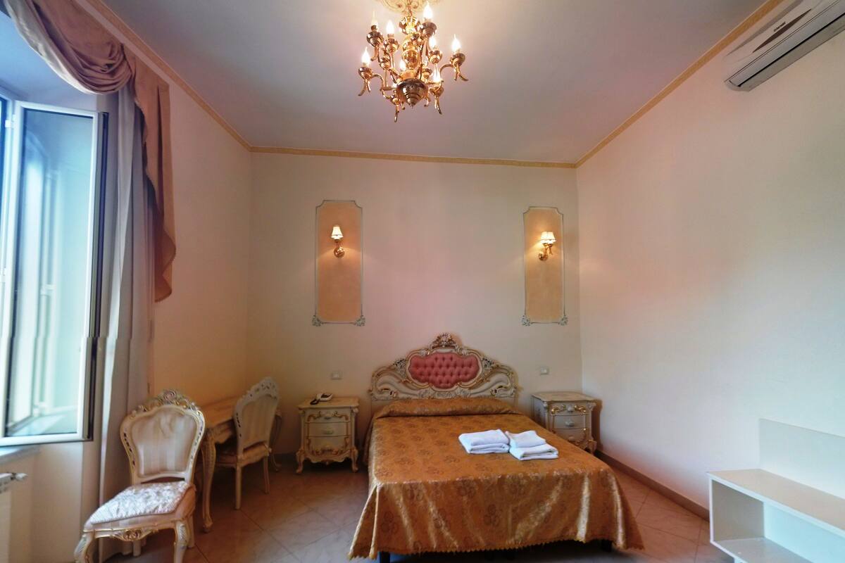A bedroom with a large bed, a chandelier, two wall sconces, a chair, a nightstand, and a radiator.
