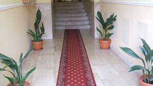 A hallway with a red patterned carpet, potted plants on either side, and a staircase in the background. - Imperial Rooms (Rome)