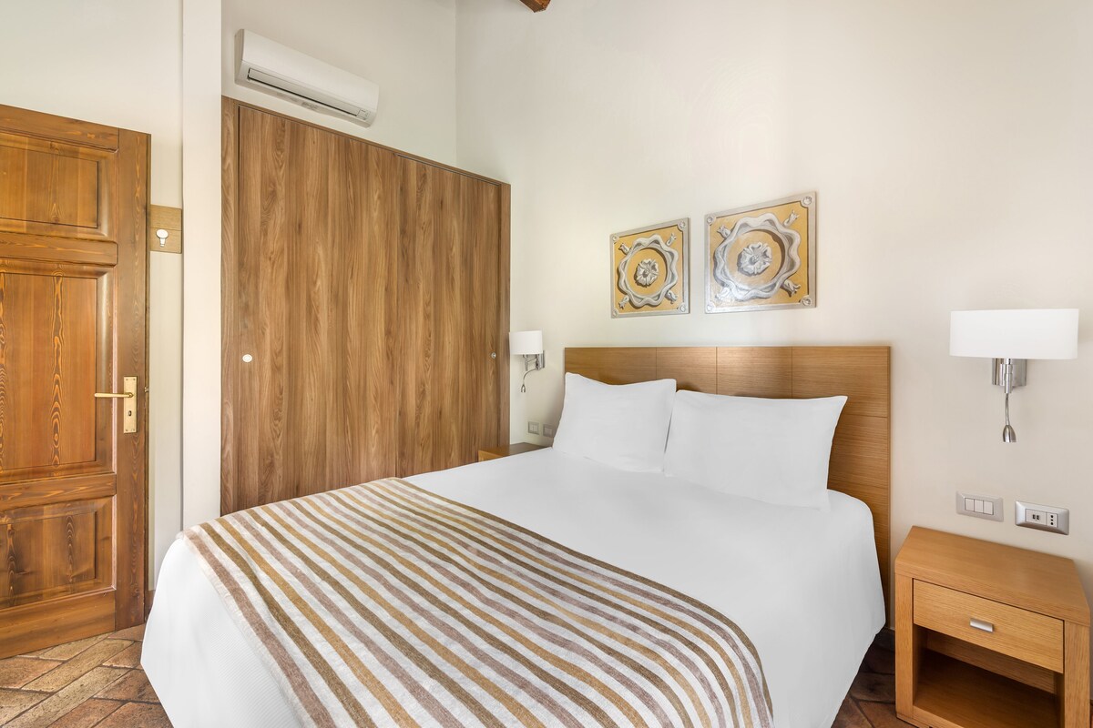 A hotel room with a large bed, wooden wardrobe, two wall-mounted lamps, and two decorative wall pieces.