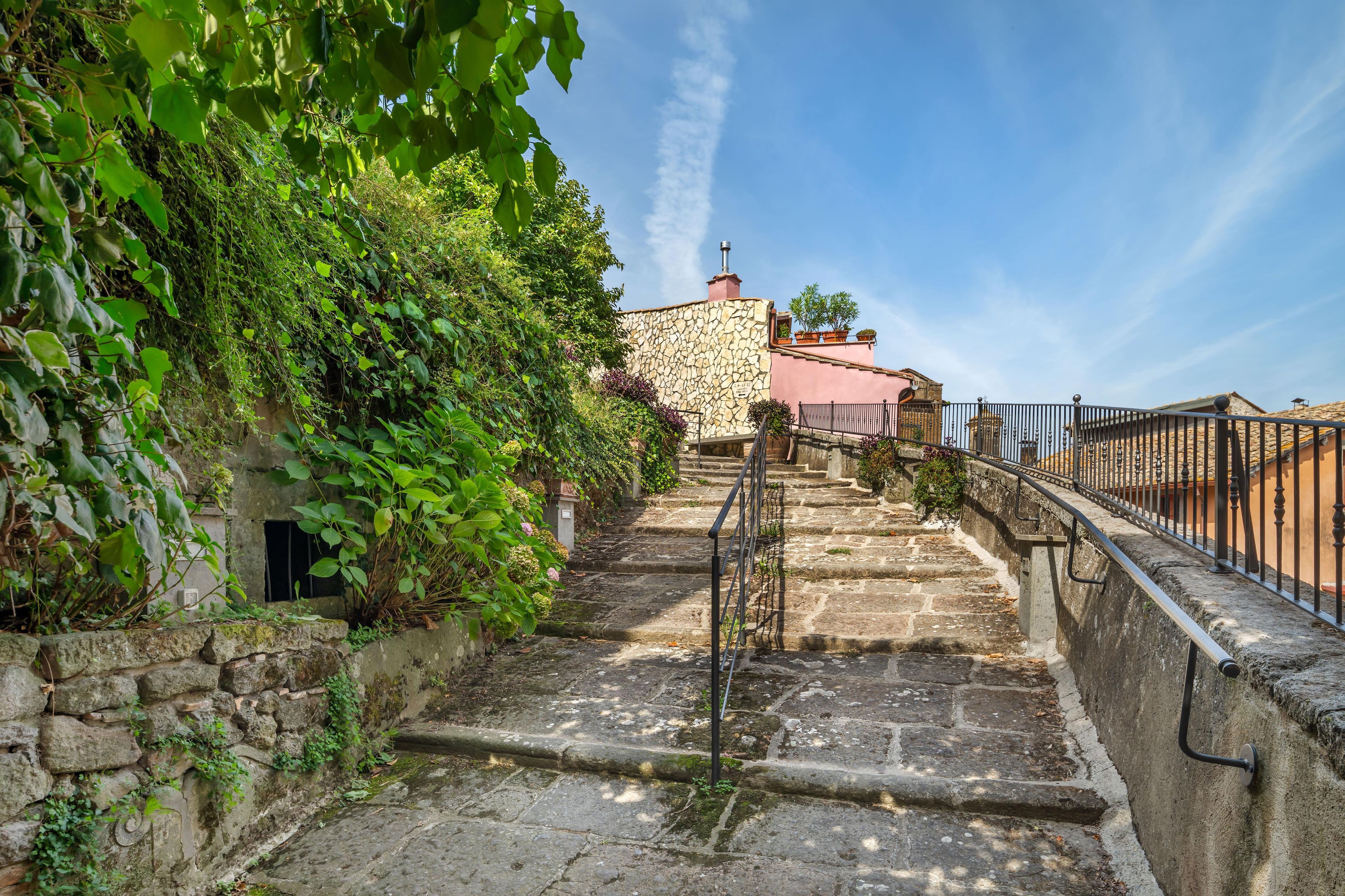 A stone staircase leading to a building with a stone facade and a pink roof, surrounded by greenery.