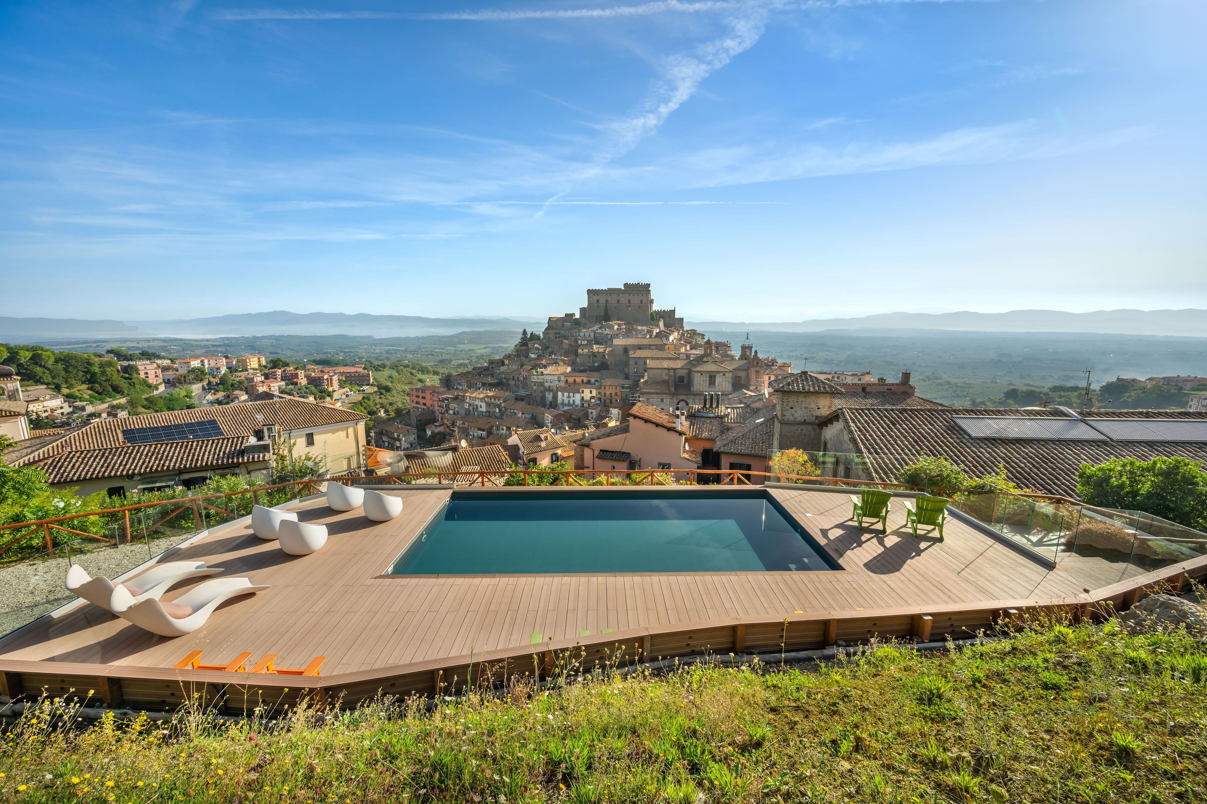A rooftop pool with lounge chairs, a view of a historic town with a castle, and surrounding greenery.