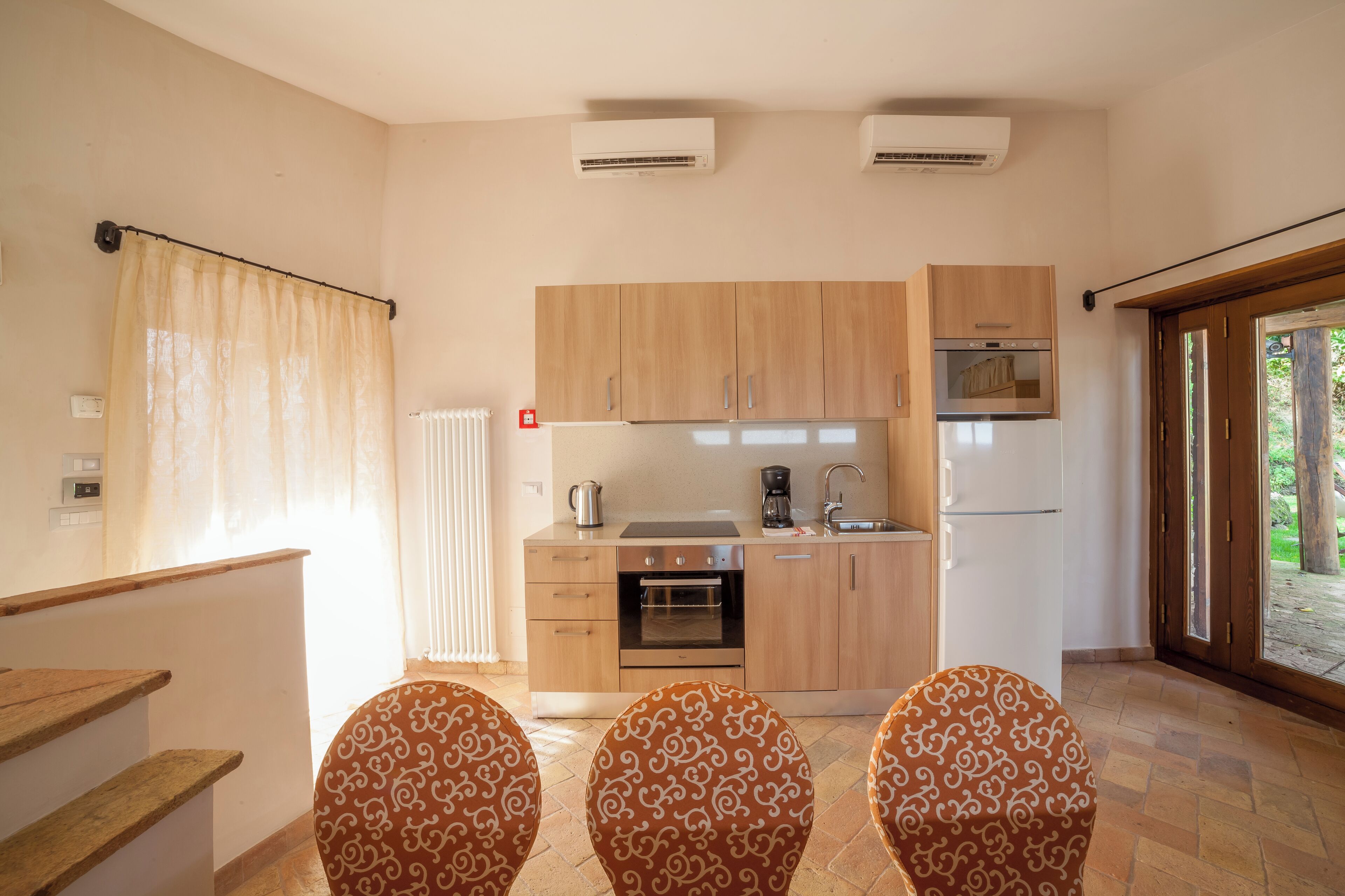 Una cucina compatta con mobili in legno, forno integrato e frigorifero. Sono presenti due sedie decorate e una finestra con tende.