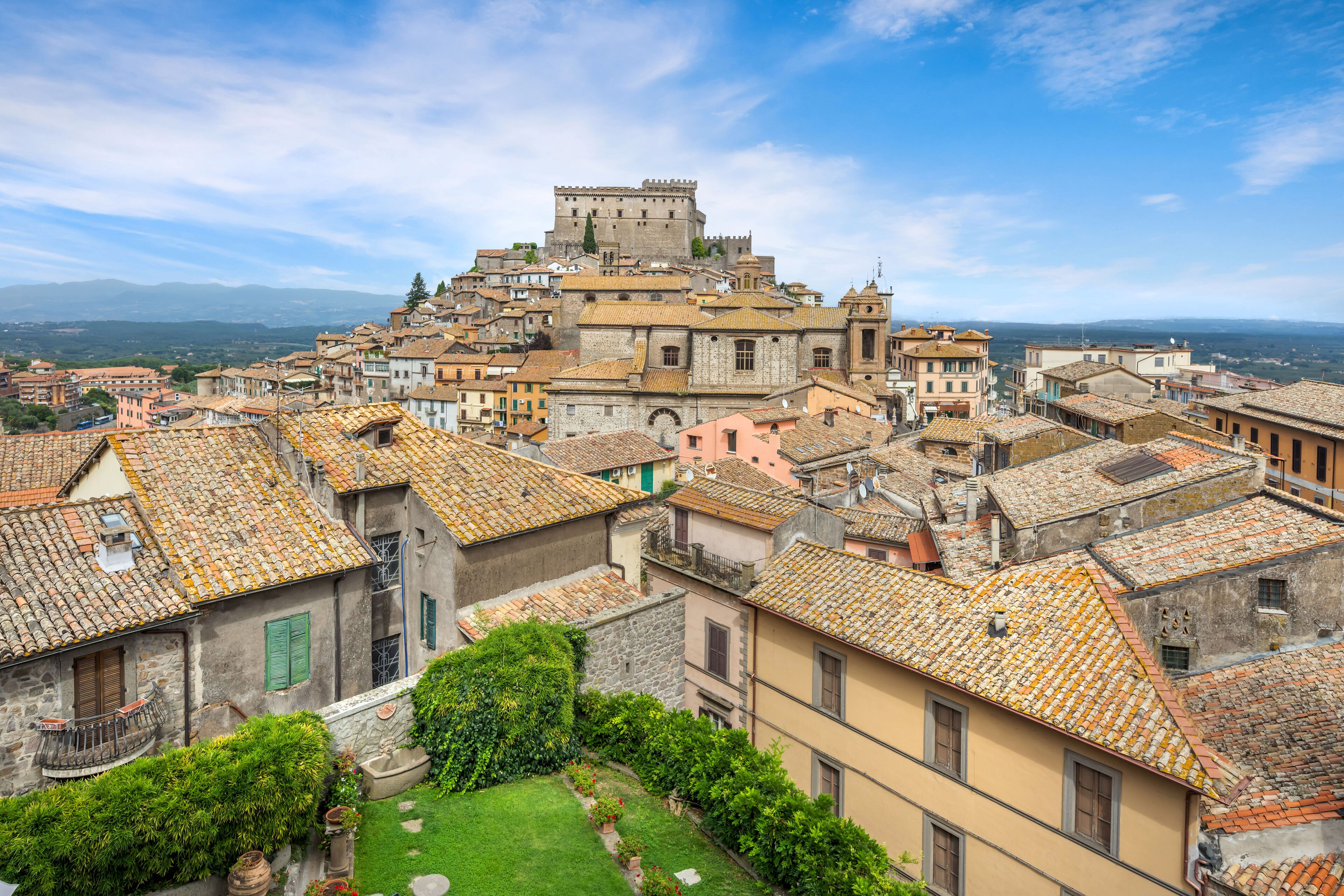 Une ville chargée d’histoire, avec un château imposant qui domine la colline, des bâtiments aux toits de tuiles rouges et une cour verdoyante et luxuriante.